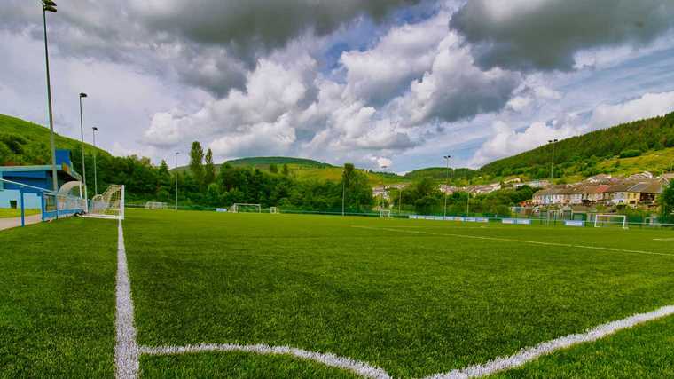 Natural grass vs synthetic turf comparison showing a green athletic field surrounded by hills and cloudy skies.
